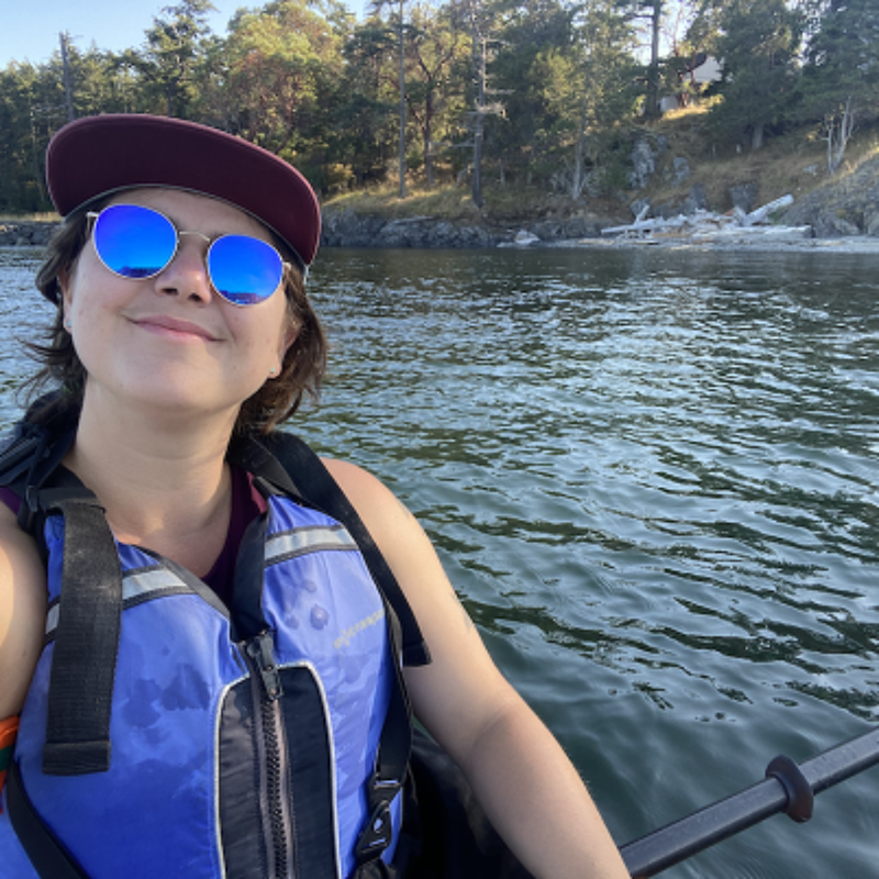 The image shows a woman in a kayak on the water. She is wearing sunglasses, a hat, and a life vest. She is smiling at the camera. The background includes trees and a shoreline. The lighting suggests it is daytime.